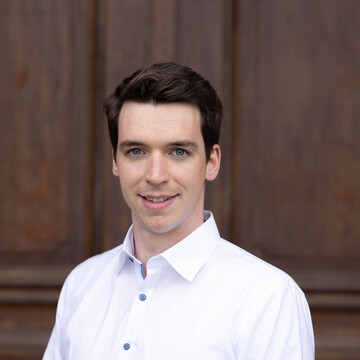Lukas Mahler. Er trägt ein weißes Hemd und steht vor einer dunkelbraunen Holztür.