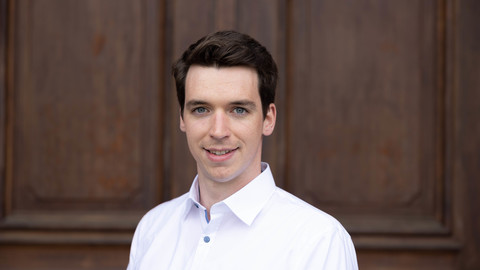 Lukas Mahler. He is wearing a white shirt and is standing in front of a brown door.