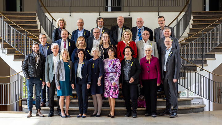 Gruppenfoto von Erwachsenen in formeller Kleidung, die vor einer Treppe Ostflügel des Mannheimer Schlosses posieren. Einige tragen gelbe Schleifen als Anstecker.