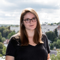 Jana Schindin hat glatte braune Haare und eine Brille. Sie trägt ein schwarzes T-Shirt.