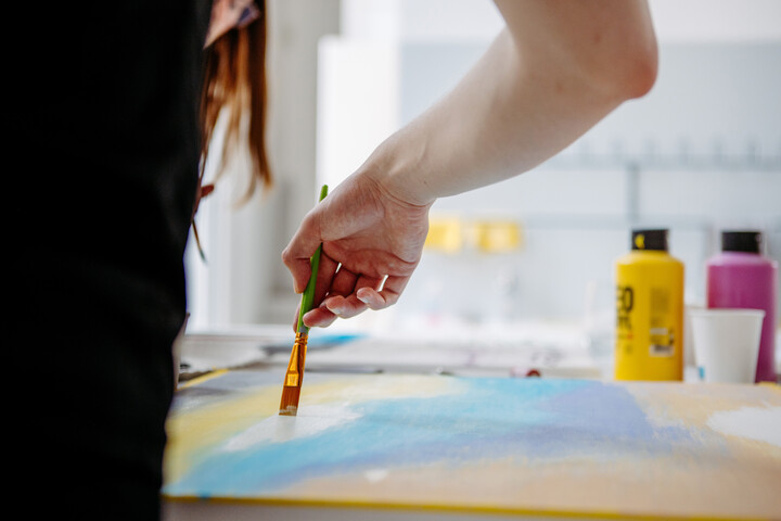 Person malt mit einem Pinsel auf einer Leinwand, im Hintergrund sind bunte Farbtuben zu sehen.