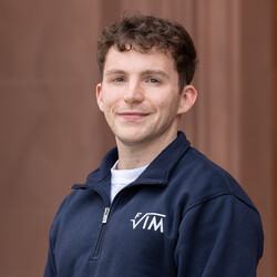 Yannick, mit kurzen hellbraunen Haaren trägt einen dunkelblauen Pullover der Fachschaft und lächelt freundlich in die Kamera