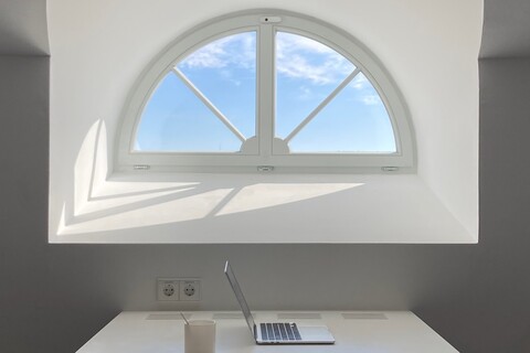 Vor einem kleinen Fenster ist ein Arbeitstisch auf dem ein aufgeklappter Laptop und eine Tasse liegen.