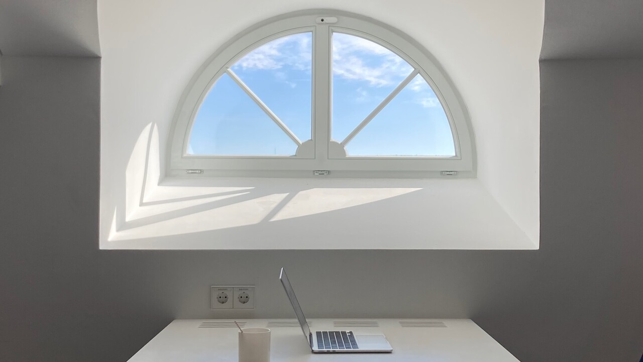 Vor einem kleinen Fenster ist ein Arbeitstisch auf dem ein aufgeklappter Laptop und eine Tasse liegen.