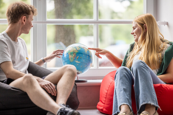 Zwei Studierende sitzen in einem Sitzsack und zeigen auf den, zwischen ihnen stehenden, Globus. Sie lächeln dabei.