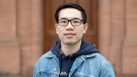 Youpeng Zhang. He is wearing a blue jeans jacket over a dark blue hoodie and is standing in front of a brown door.