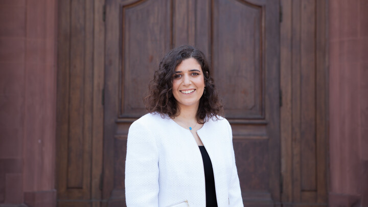 Eine lächelnde Person trägt ein schwarzes Top sowie einen weißen Blazer und steht vor einer Holztüre. Die Person heißt Amina Elbarbary. 
