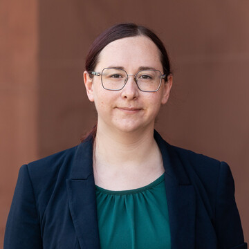 Elke Baumann hat dunkelrote Haare. Sie trägt eine Brille und ein dunkelgrünes Oberteil mit einem dunkelblauen Blazer darüber.