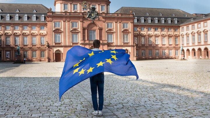 Eine Person läuft von der Kamera weg über den Ehrenhof und lässt eine EU-Flagge über den Rücken wehen. Im Hintergrund ist das Mannheimer Barockschloss zu sehen.
