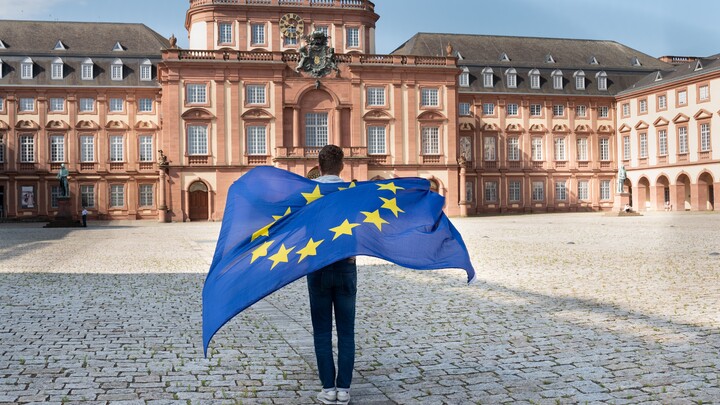 Eine Person läuft von der Kamera weg über den Ehrenhof und lässt eine EU-Flagge über den Rücken wehen. Im Hintergrund ist das Mannheimer Barockschloss zu sehen.