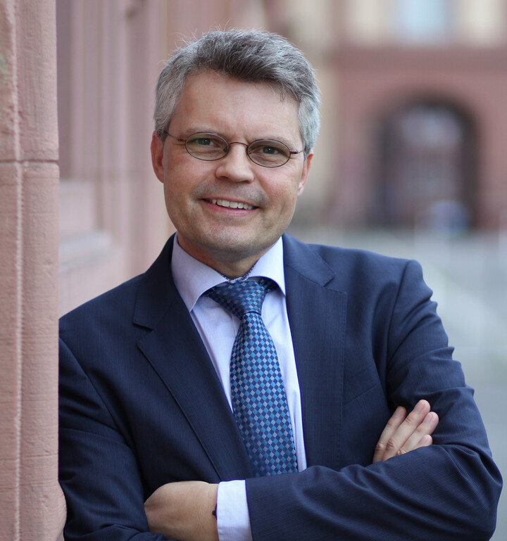 Prof. Dr. Ralf Müller-Terpitz mit ovaler Brille in dunkelblauem Anzug und blau-weißer Krawatte mit verschränkten Armen an das Schloss lehnend