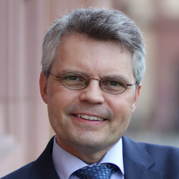 Prof. Dr. Ralf Müller-Terpitz mit ovaler Brille in dunkelblauem Anzug und blau-weißer Krawatte mit verschränkten Armen an das Schloss lehnend