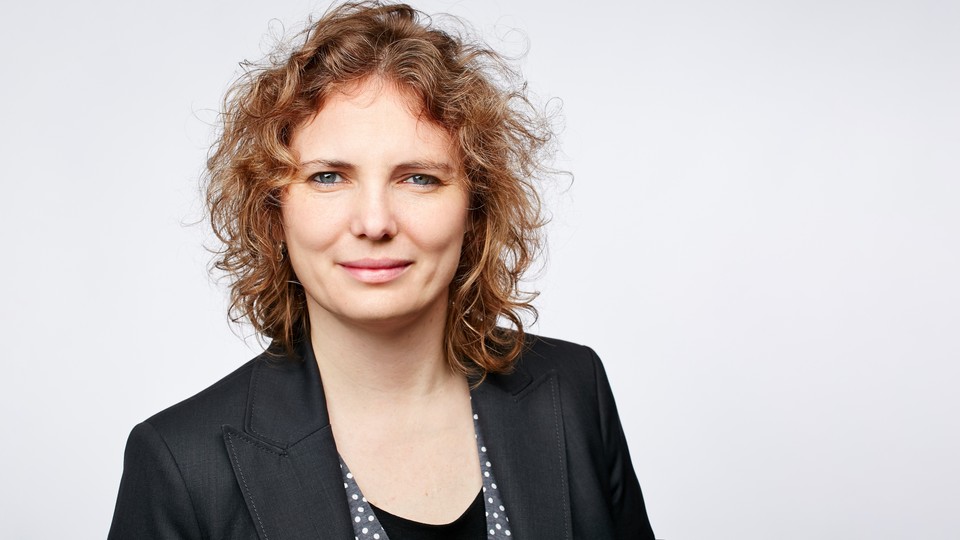 Portrait von Johanna Gereke. Sie trägt einen schwarzen Blazer und steht vor einer hellgrauen Wand.