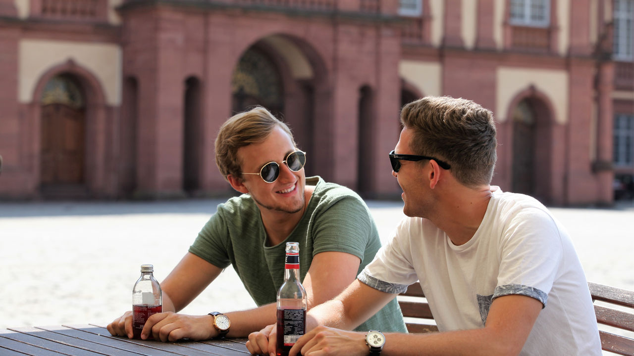 Zwei Studenten lächeln sich an während sie mit ihren Getränken an einem Tisch im Ehrenhof des Mannheimer Schlosses sitzen. Sie tragen T-Shirts und Sonnenbrillen.
