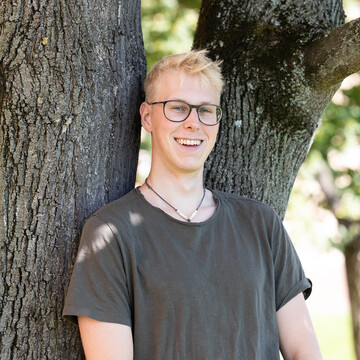  Renke Meyer. Er trägt eine Brille und ein dunkelgrünes T-Shirt, dabei lehnt er lachend an einem Baum.