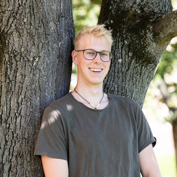 Renke Meyer. Er trägt eine Brille und ein dunkelgrünes T-Shirt, dabei lehnt er lachend an einem Baum.
