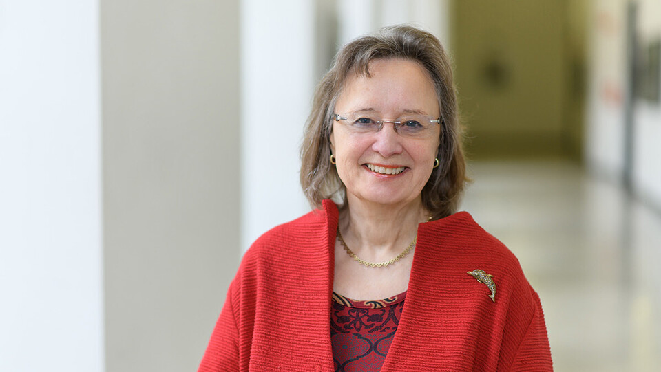Portrait von Rosemarie Tracy. Sie trägt eine rote Strickjacke mit einer goldenen Fischbrosche. 