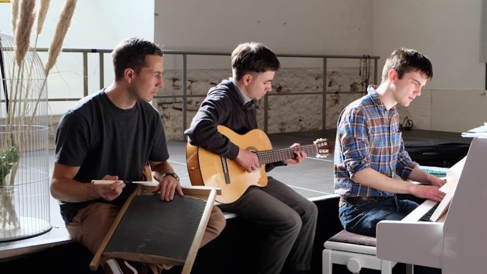 Drei Personen sitzen nenbeneinander und musizieren zusammen. Eine Person sitzt an einem Klavier, eine andere spielt Gitarre.