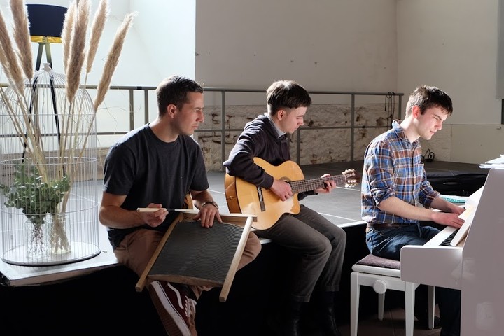 Drei Personen sitzen nenbeneinander und musizieren zusammen. Eine Person sitzt an einem Klavier, eine andere spielt Gitarre.