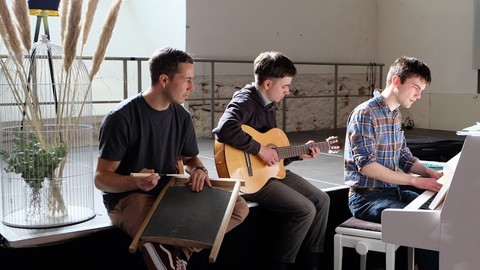 Drei Personen sitzen nenbeneinander und musizieren zusammen. Eine Person sitzt an einem Klavier, eine andere spielt Gitarre.