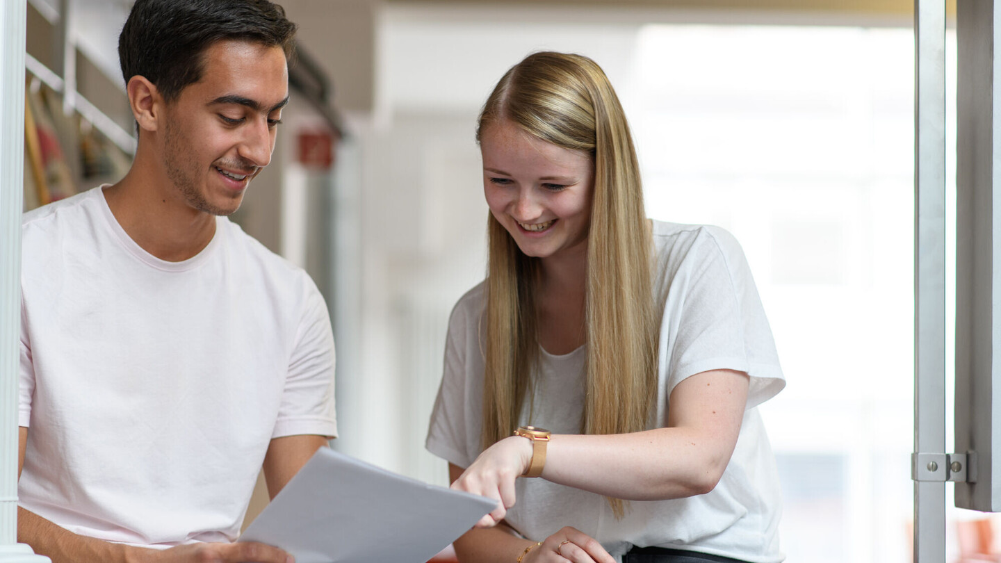 Zwei Personen stehen beieinander und sprechen über einen Text, den die eine Person ausgedruckt in den Händen hält.