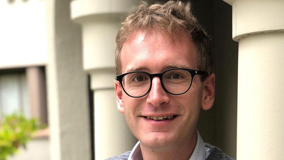 Adrian Daub lehnt mit verschränkten Armen an einer Säule und lächelt in die Kamera.