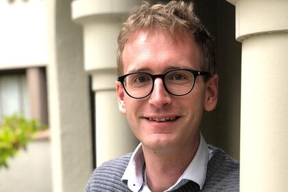 Adrian Daub lehnt mit verschränkten Armen an einer Säule und lächelt in die Kamera.