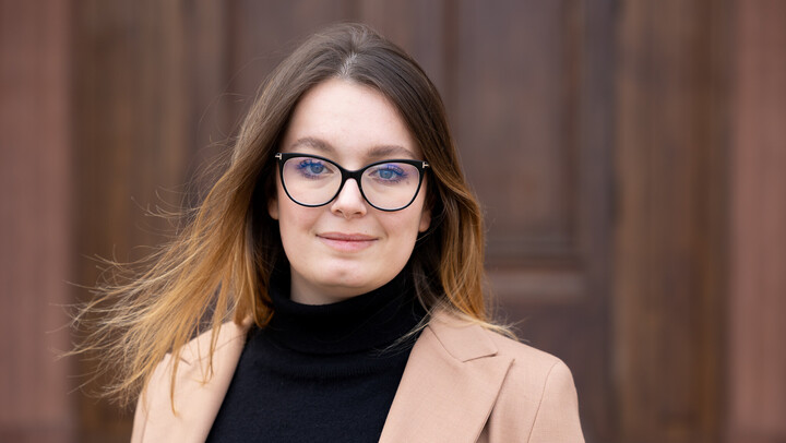 Chiara Malavasi. She is wearing a beige jacket over a black sweater and is standing in front of brown door.