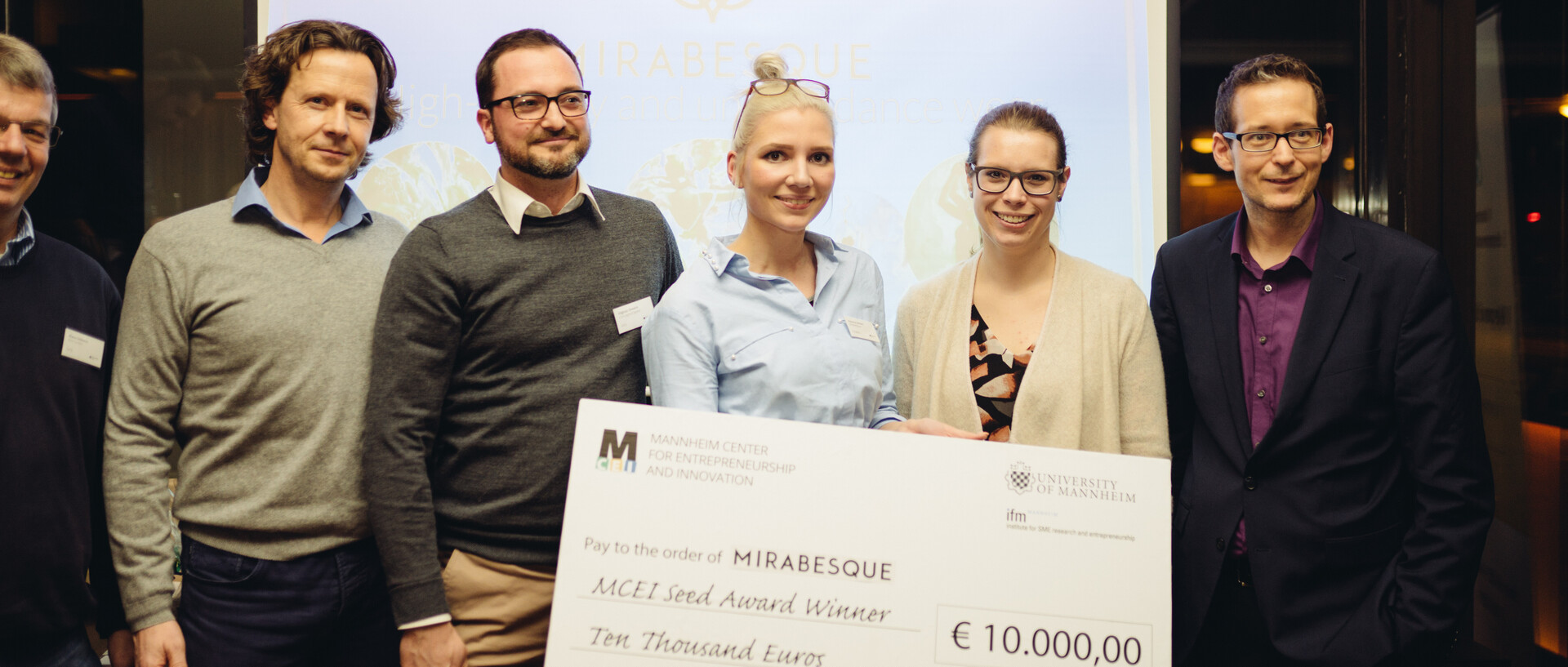 Sechs Personen stehen vor einer Leinwand und halten ein Schild mit einem Scheck des MCEIs über 10.000€ in der Hand.