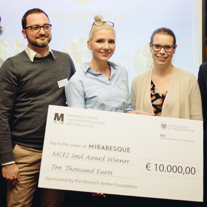 Sechs Personen stehen vor einer Leinwand und halten ein Schild mit einem Scheck des MCEIs über 10.000€ in der Hand.