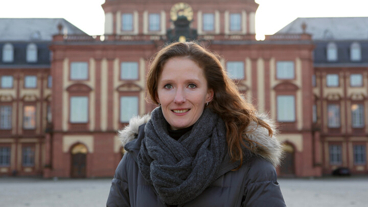 Portrait von Karin Hoisl. Sie trägt eine Winterjacke und einen grauen Schal und steht auf dem Ehrenhof vor dem Schloss. 