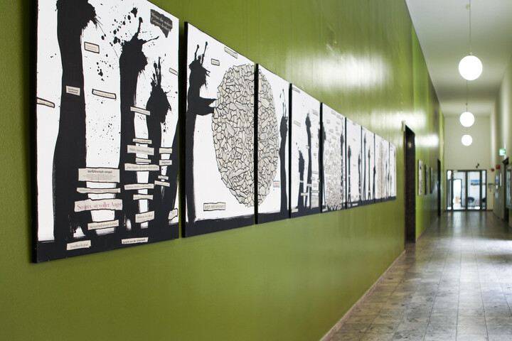 In einem Flur mit grüner Wand hängen Kunstwerke bestehend aus Zeitungscollagen und schwarzen Farbklecksen.