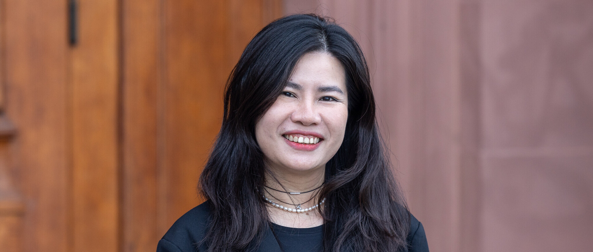 Porträt von Thuy Nguyen. Sie steht vor einem braunen Hintergrund, trägt ein schwarzes Jackett und lächelt in die Kamera.