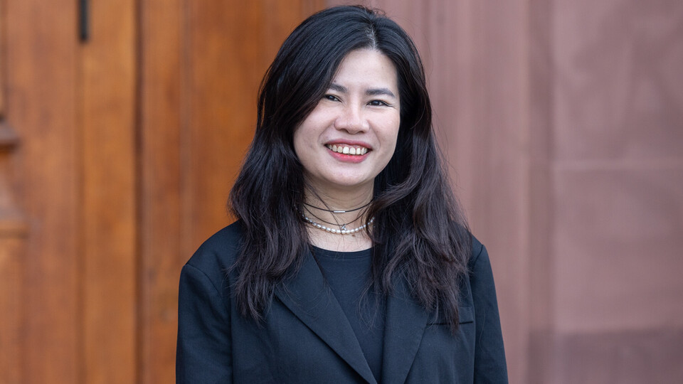 Porträt von Thuy Nguyen. Sie steht vor einem braunen Hintergrund, trägt ein schwarzes Jackett und lächelt in die Kamera.