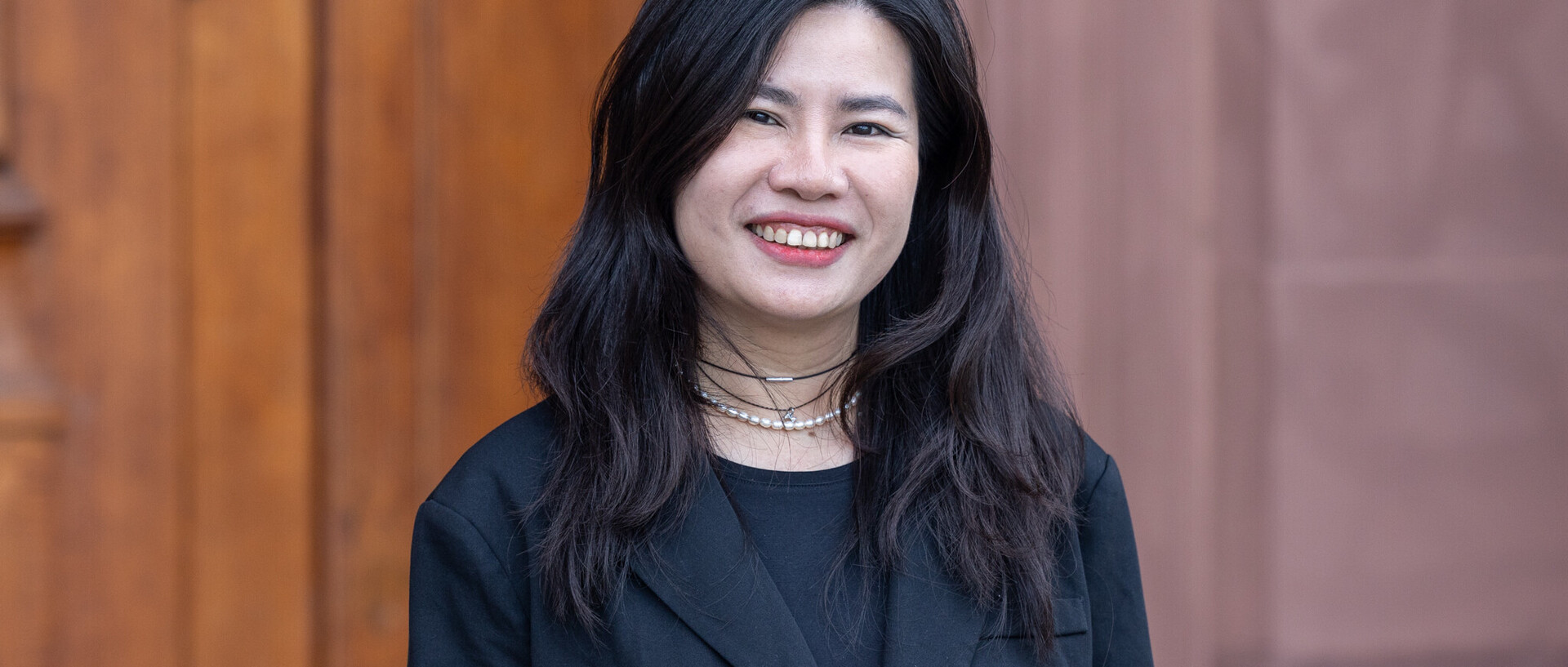 Porträt von Thuy Nguyen. Sie steht vor einem braunen Hintergrund, trägt ein schwarzes Jackett und lächelt in die Kamera.