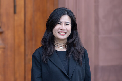 Porträt von Thuy Nguyen. Sie steht vor einem braunen Hintergrund, trägt ein schwarzes Jackett und lächelt in die Kamera.