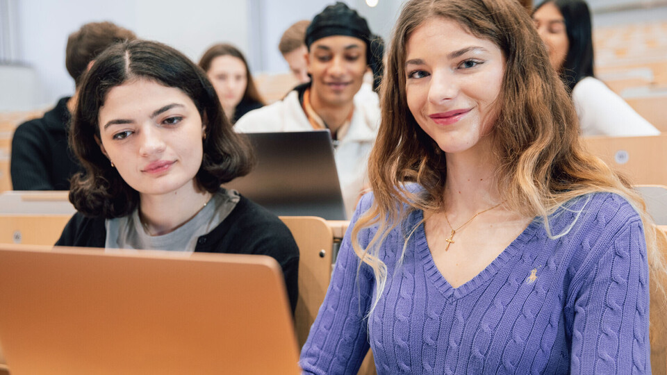 Studierende sitzen in einem Hörsaal. Zwei Studentinnen sind im Fokus. Sie schauen auf einen Laptop. 