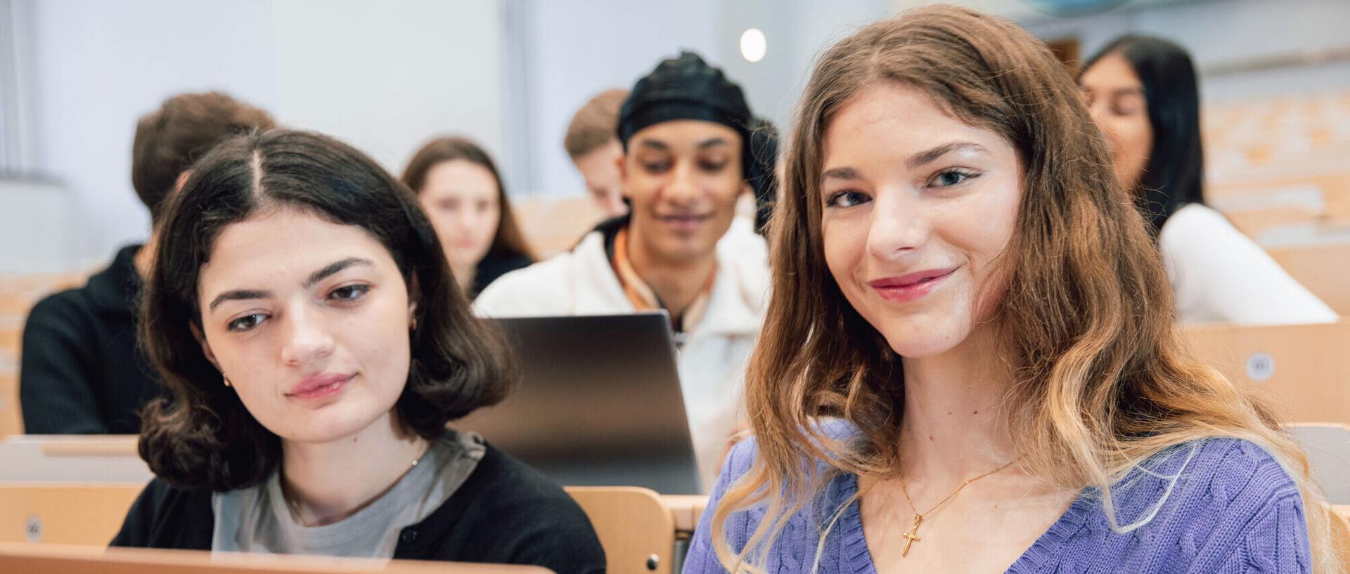 Studierende sitzen in einem Hörsaal. Zwei Studentinnen sind im Fokus. Sie schauen auf einen Laptop.