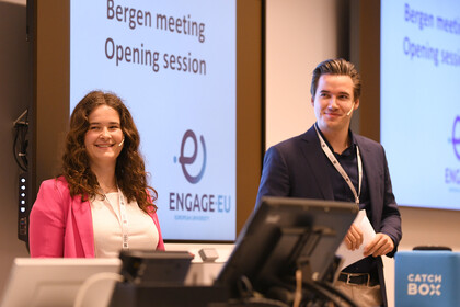 Zwei Personen in schicker Kleidung stehen nebeneinander. Auf die Wand hinter ihnen wird das Logo von Engage.Eu sowie die Schrift "Bergen meeting opening session" projiziert.