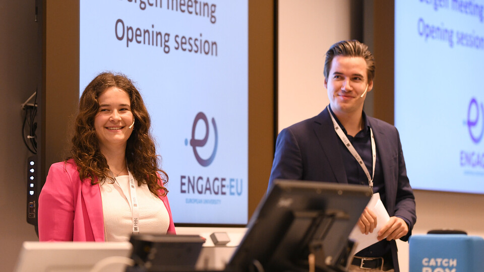 Zwei Personen in schicker Kleidung stehen nebeneinander. Auf die Wand hinter ihnen wird das Logo von Engage.Eu sowie die Schrift "Bergen meeting opening session" projiziert.
