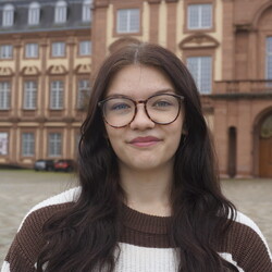 Maresa trägt lockigen Haaren und Brille und lächelt freundlich in die Kamera mit dem Schloss im Hintergrund.