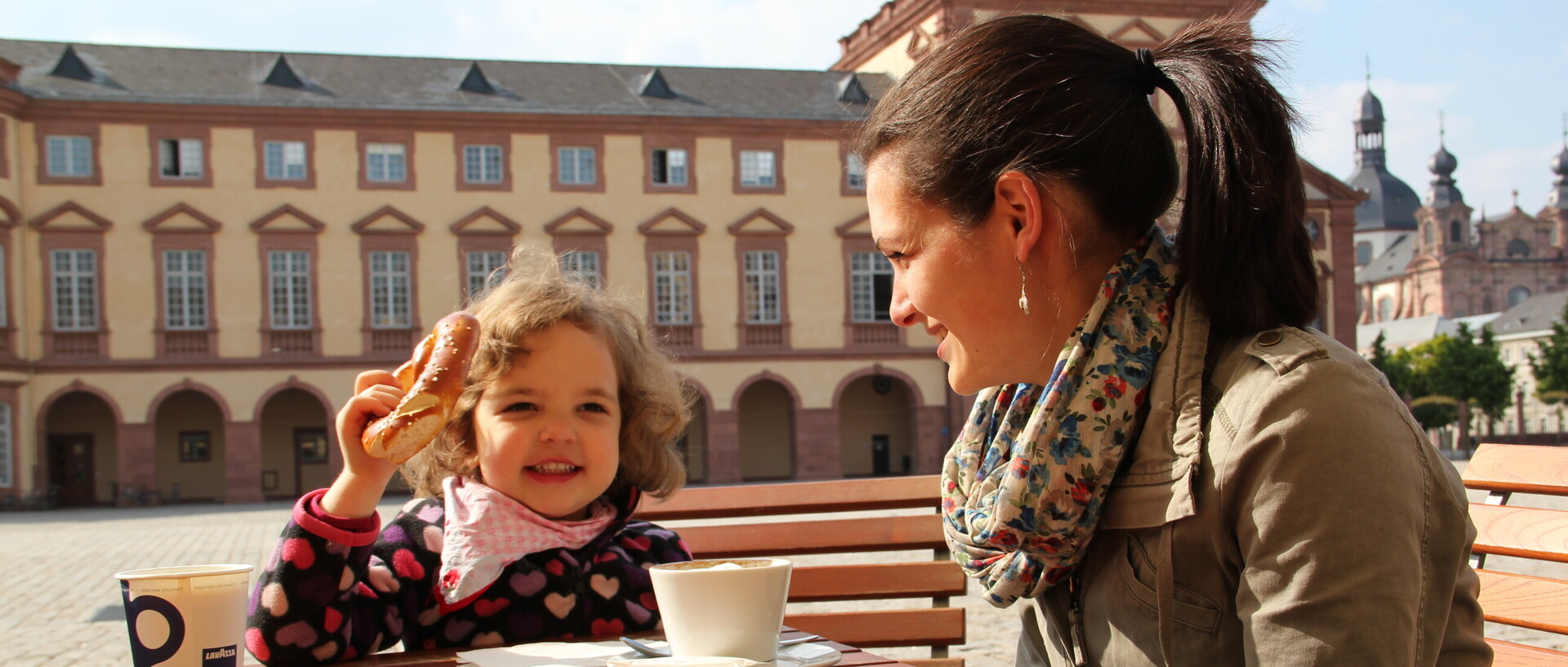 Frau mit kleinem Kind sitzt im Ehrenhof der Universität, das Kind isst eine Bretzel, die Frau trinkt Kaffee, sonniger Tag.