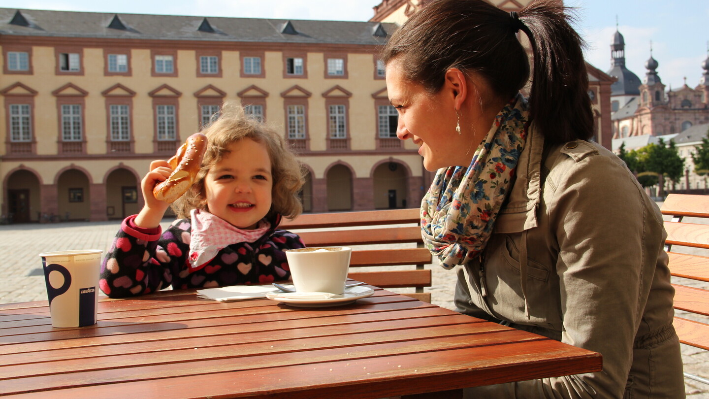 Frau mit kleinem Kind sitzt im Ehrenhof der Universität, das Kind isst eine Bretzel, die Frau trinkt Kaffee, sonniger Tag.
