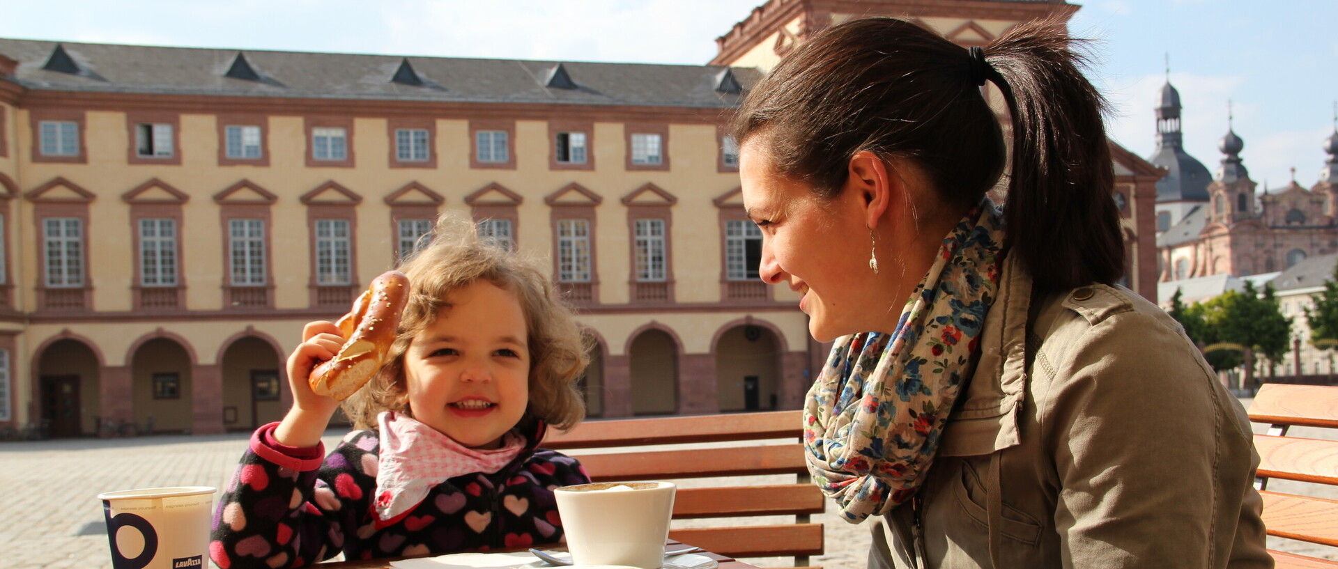 Frau mit kleinem Kind sitzt im Ehrenhof der Universität, das Kind isst eine Bretzel, die Frau trinkt Kaffee, sonniger Tag.
