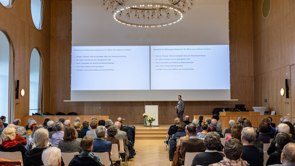 Foto von der Jubiläumsveranstaltung in der Aula. Das Publikum hört dem Redner auf dem Podium zu. Hinter ihm ist eine Präsentation an die Wand projiziert.