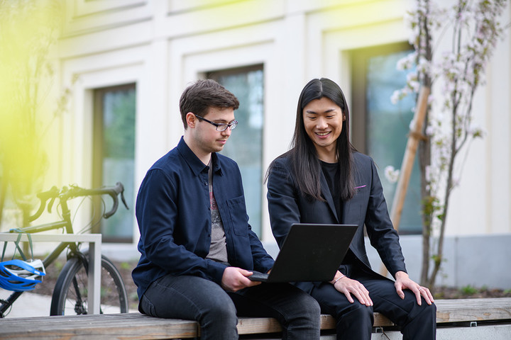 Two men are sitting on a bank in the green courtyard of the B6 building and are looking with a smile at a laptop screen. 