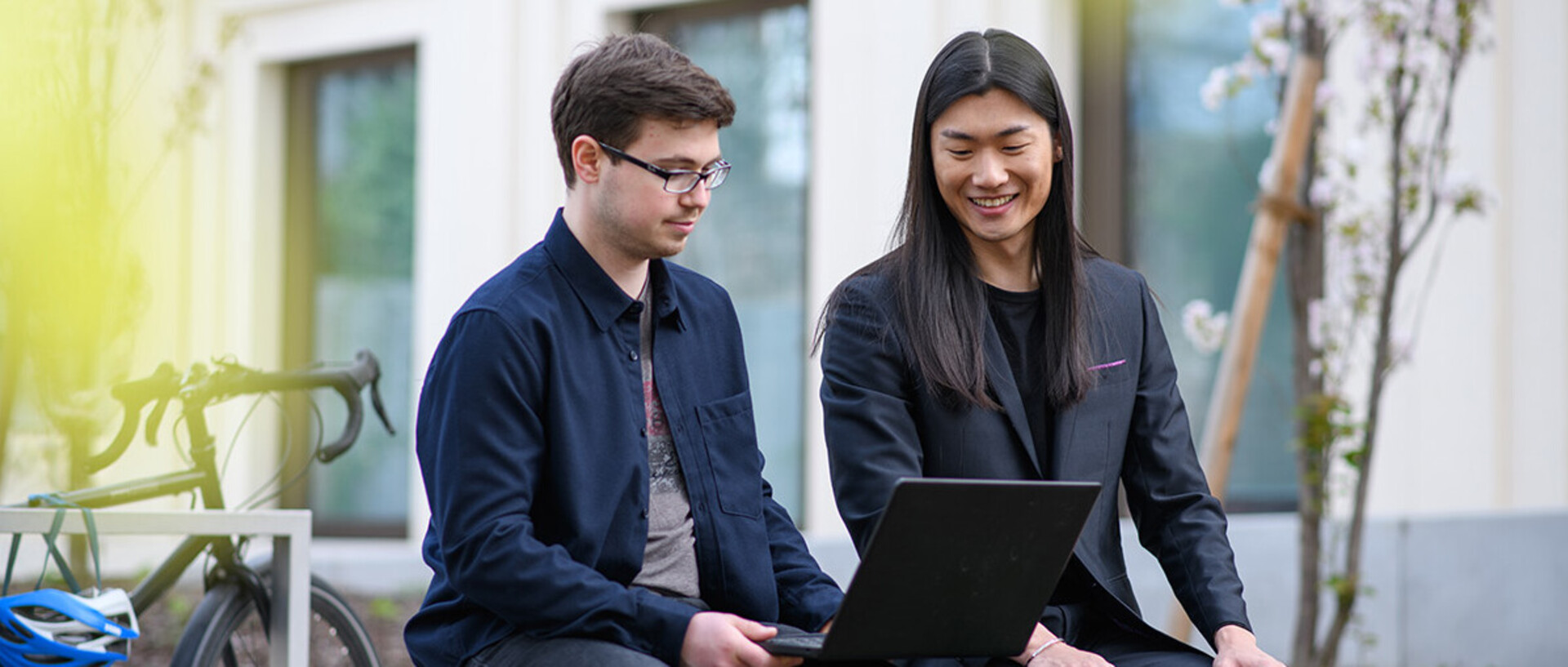 Two men are sitting on a bank in the green courtyard of the B6 building and are looking with a smile at a laptop screen.