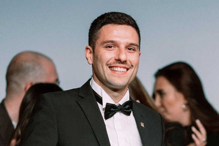 Johannes Berg ist Alumnus der Uni, er trägt einen dunklen Anzug, ein weißes Hemd und eine Fliege. Er hat dunkle kurze Haare und lächelt.