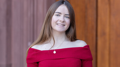 Arina Vlasova trägt einen roten Pullover und steht lächelnd vor einer braunen Holztüre. 