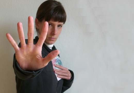Eine Frau blickt leicht nach unten in die Kamera, hält in einer Hand mehrere Geldscheine an ihre Brust und streckt den Arm und die Handfläche der anderen Hand vor ihrem Oberkörper wie als Stoppsignal aus.